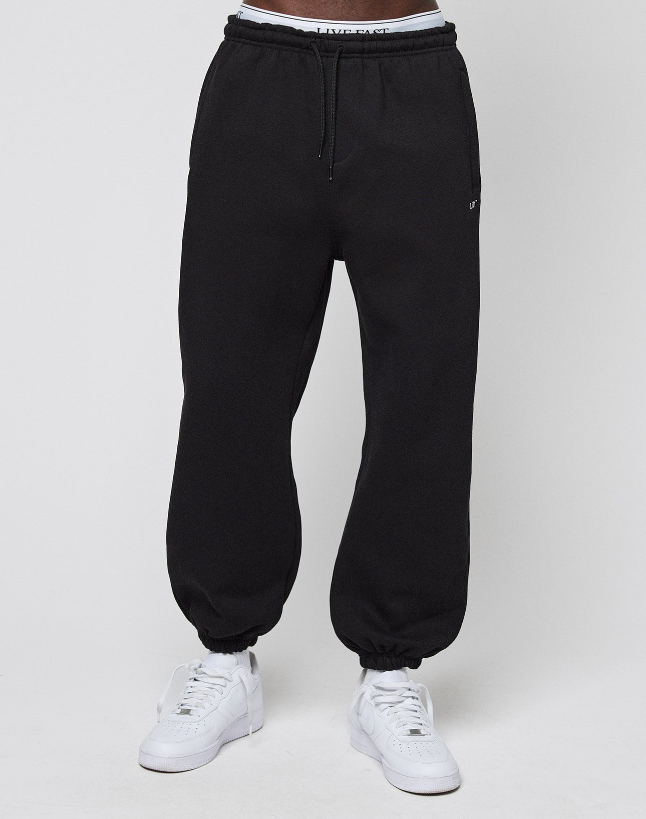 A person wears LIVE FAST DIE YOUNG Basic Loose Jogger sweatpants with an elastic waistband and cuffs, paired with white sneakers and visible white underwear, standing against a plain white background.
