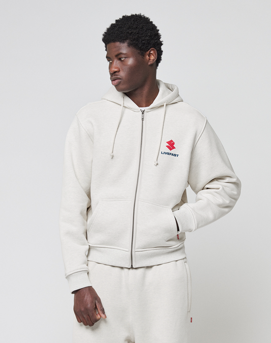 A young man wears a light beige LFDY Heavyweight Lounge Zip Hooded by LIVE FAST DIE YOUNG with matching sweatpants. The 100% cotton hoodie has a red and black LIVEFAST logo on the chest. He stands against a white background, looking to the side.