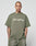 A person with a shaved head and visible tattoos wears the Basic +49 Tee by LIVE FAST DIE YOUNG, an oversized olive-green 100% Baumwolle shirt with “+49LiveFast,” plus matching shorts, against a plain light background.