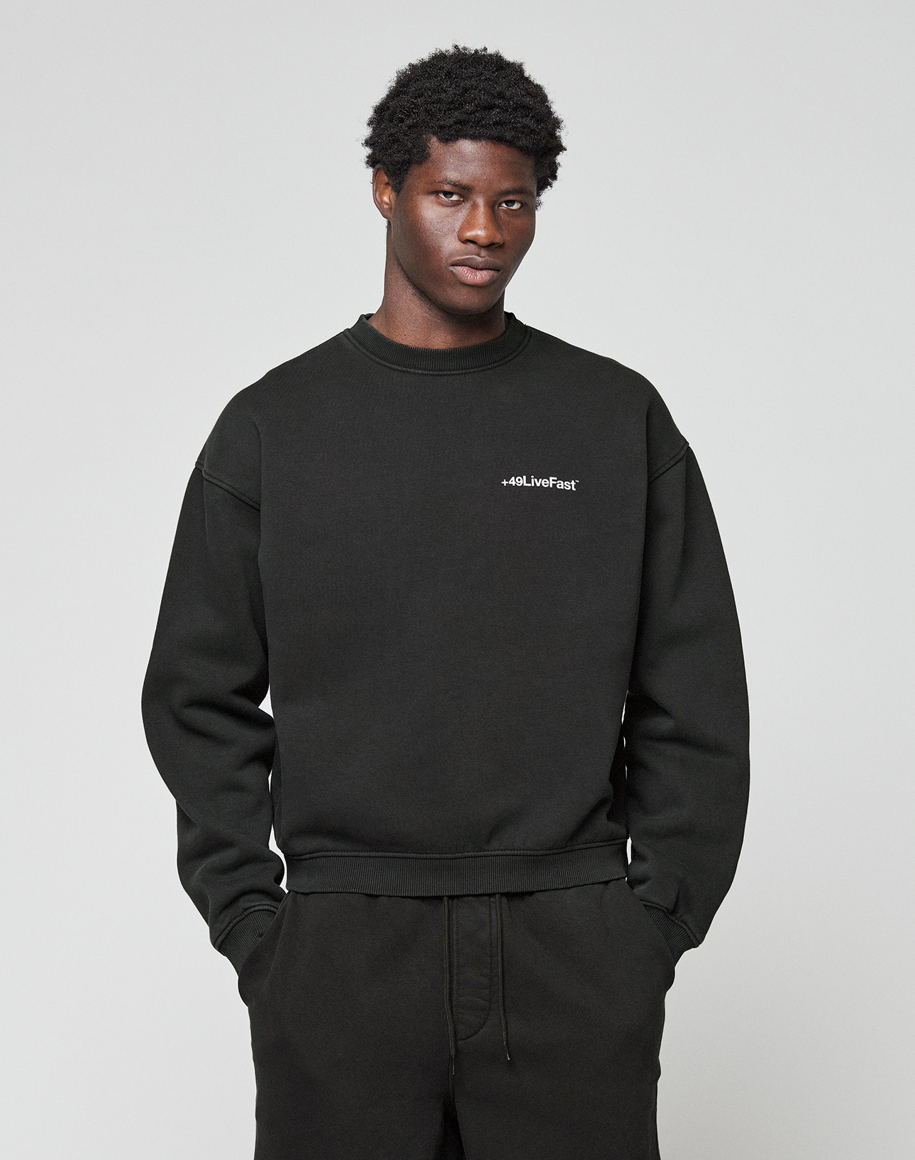 A man with short curly hair wears the LIVE FAST DIE YOUNG Basic +49 Chest Sweater in black, featuring a small white chest print, along with matching pants, and stands against a plain light gray background.