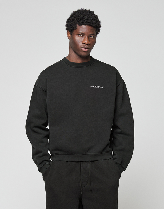 A man with short curly hair wears the LIVE FAST DIE YOUNG Basic +49 Chest Sweater in black, featuring a small white chest print, along with matching pants, and stands against a plain light gray background.