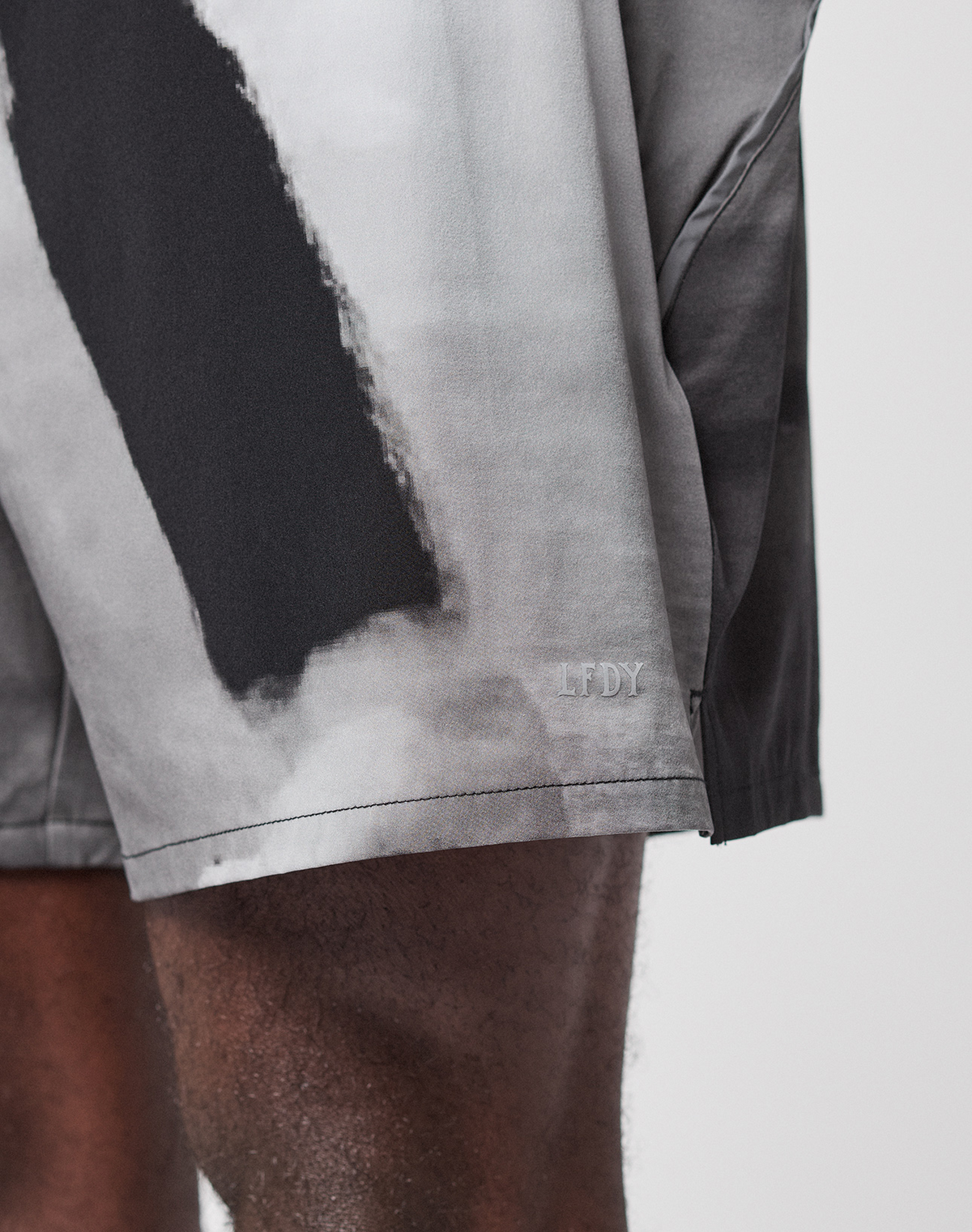 Close-up of a person's lower body wearing LIVE FAST DIE YOUNG's LFDY Performance Shorts in gray and black with a small embroidered LFDY near the hem, made from lightweight material. Background is plain and light-colored.