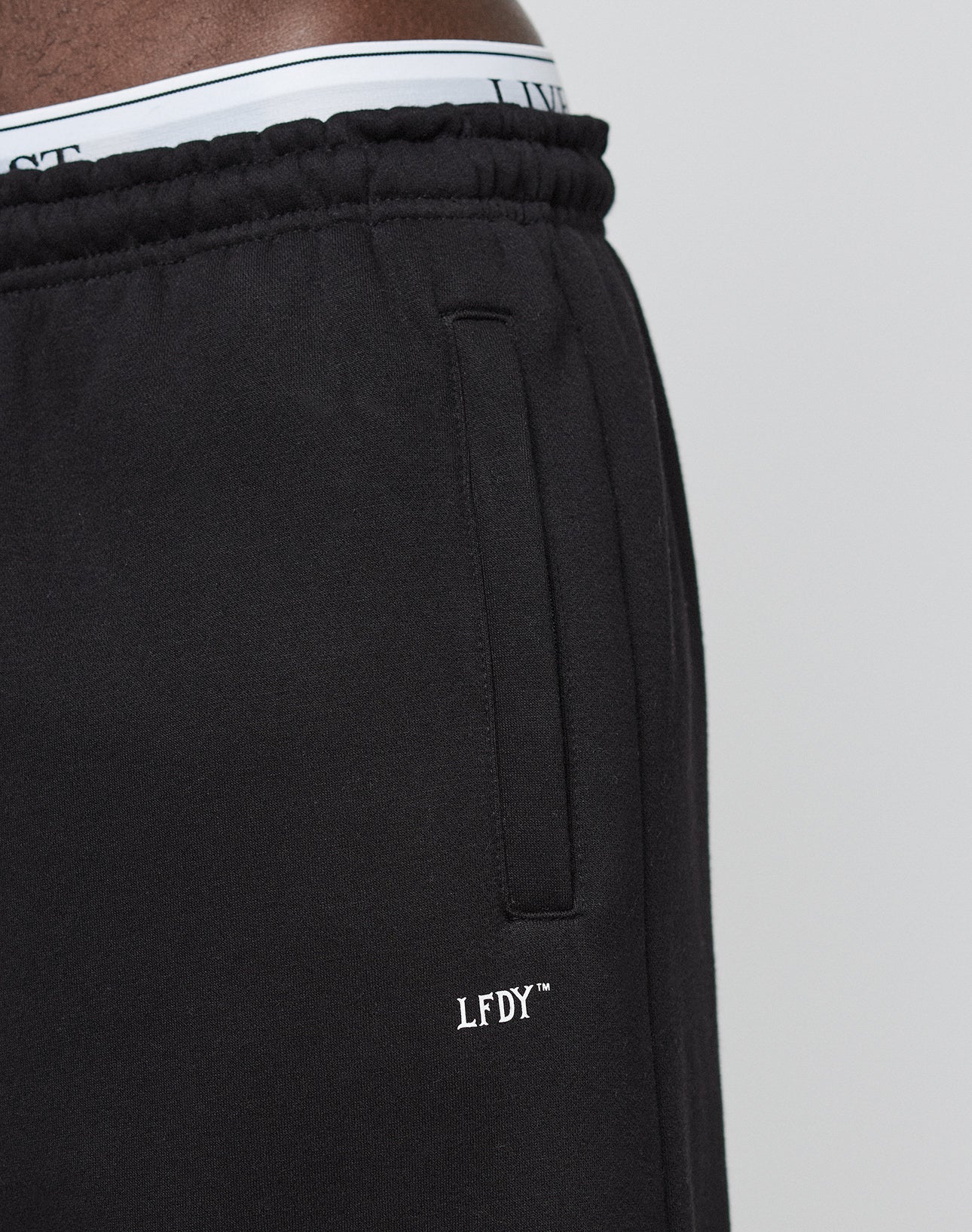 Close-up of a person wearing LIVE FAST DIE YOUNGs Basic Loose Jogger, featuring a black color, white LFDY™ logo by the pocket, elastic waistband, loose fit, and white underwear slightly visible above the pants.
