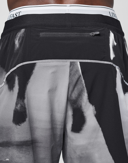 A close-up of the back of a person wearing LFDY Performance Shorts by LIVE FAST DIE YOUNG, featuring black and white abstract or animal print, a zipper pocket, "LIVE FAST" on the waistband, and a reflective logo print.