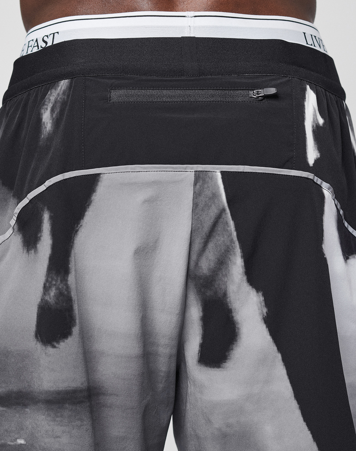 A close-up of the back of a person wearing LFDY Performance Shorts by LIVE FAST DIE YOUNG, featuring black and white abstract or animal print, a zipper pocket, "LIVE FAST" on the waistband, and a reflective logo print.