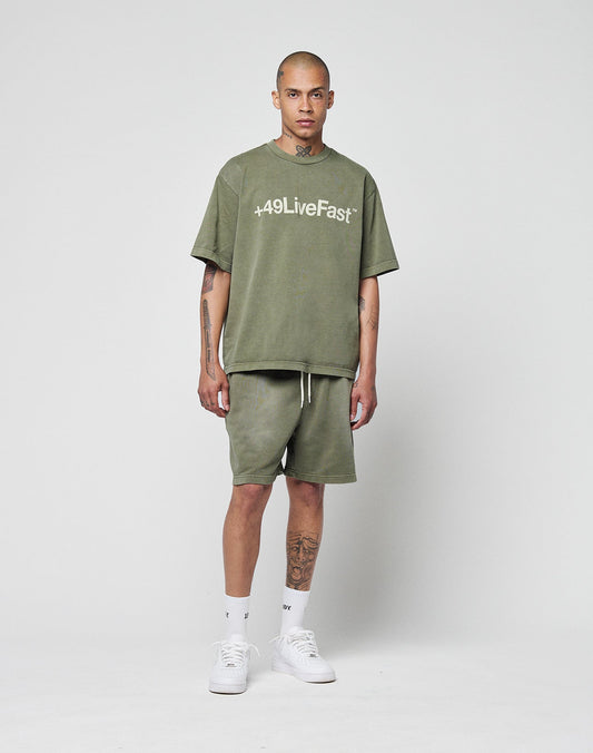 A tattooed person with a neutral expression stands against a white background, wearing olive green LIVE FAST DIE YOUNG Basic Sweat Shorts with an elastic waistband, an olive “+49LiveFast” tee, white socks, and white sneakers.