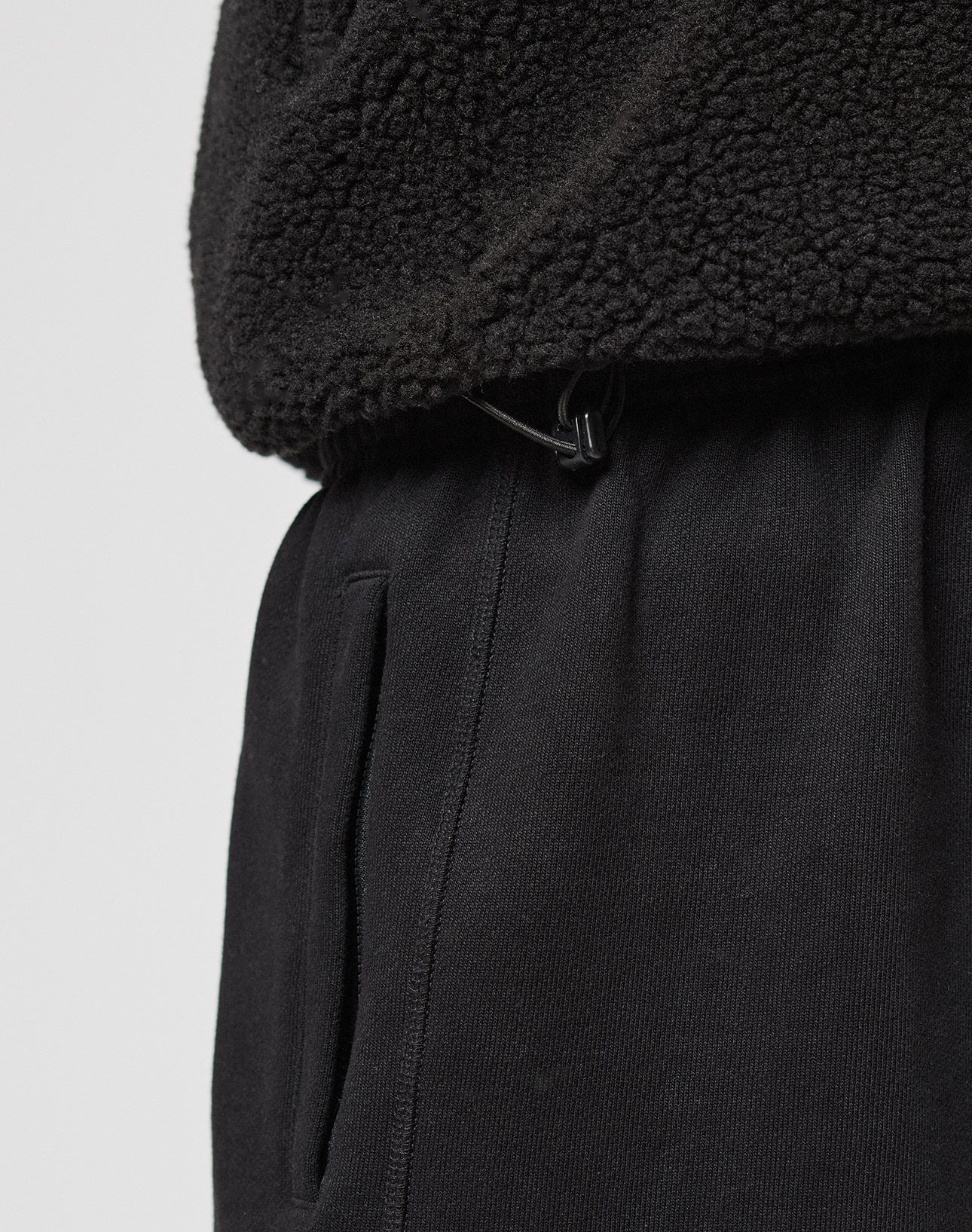 Close-up of a person wearing the black LFDY Teddy Jacket by LIVE FAST DIE YOUNG with raglan sleeves and an adjustable waist, styled with black sweatpants featuring a side pocket, set against a plain light background.