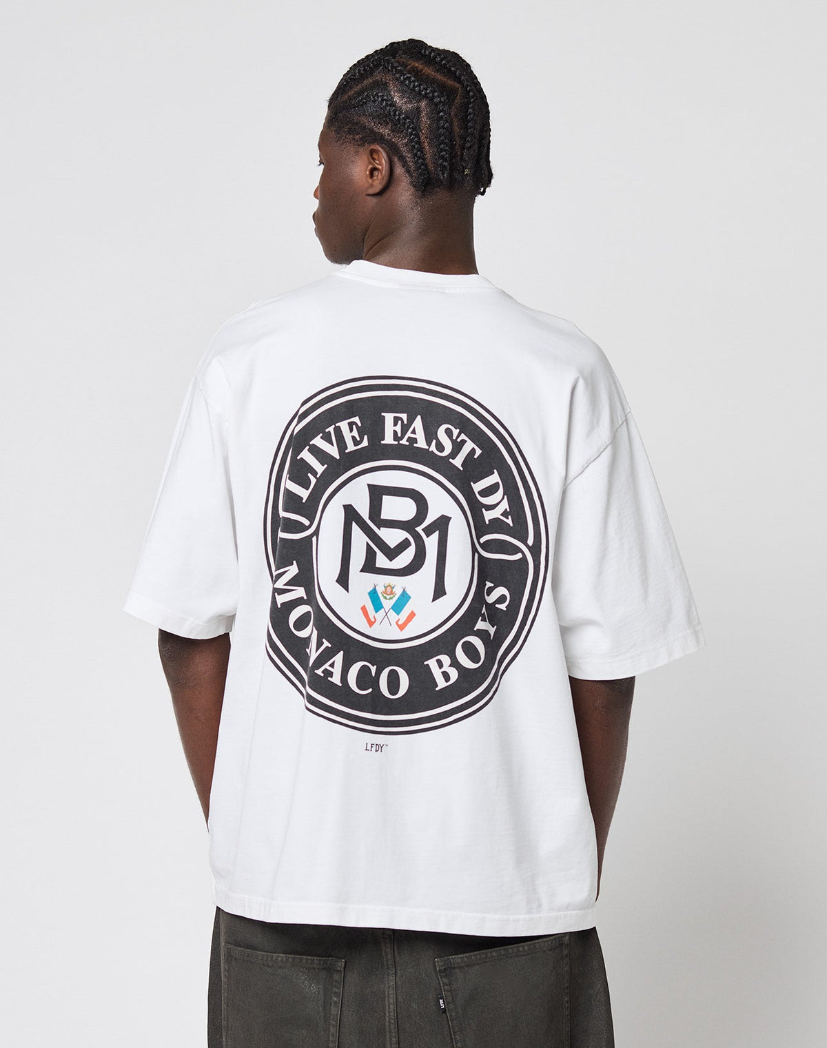 A person with braided hair faces away, wearing the LFDY Monaco Boys Tee by LIVE FAST DIE YOUNG—an oversized white 100% Baumwolle T-shirt with a large circular LIVE FAST D1 MONACO BOYS back logo and a small central emblem.