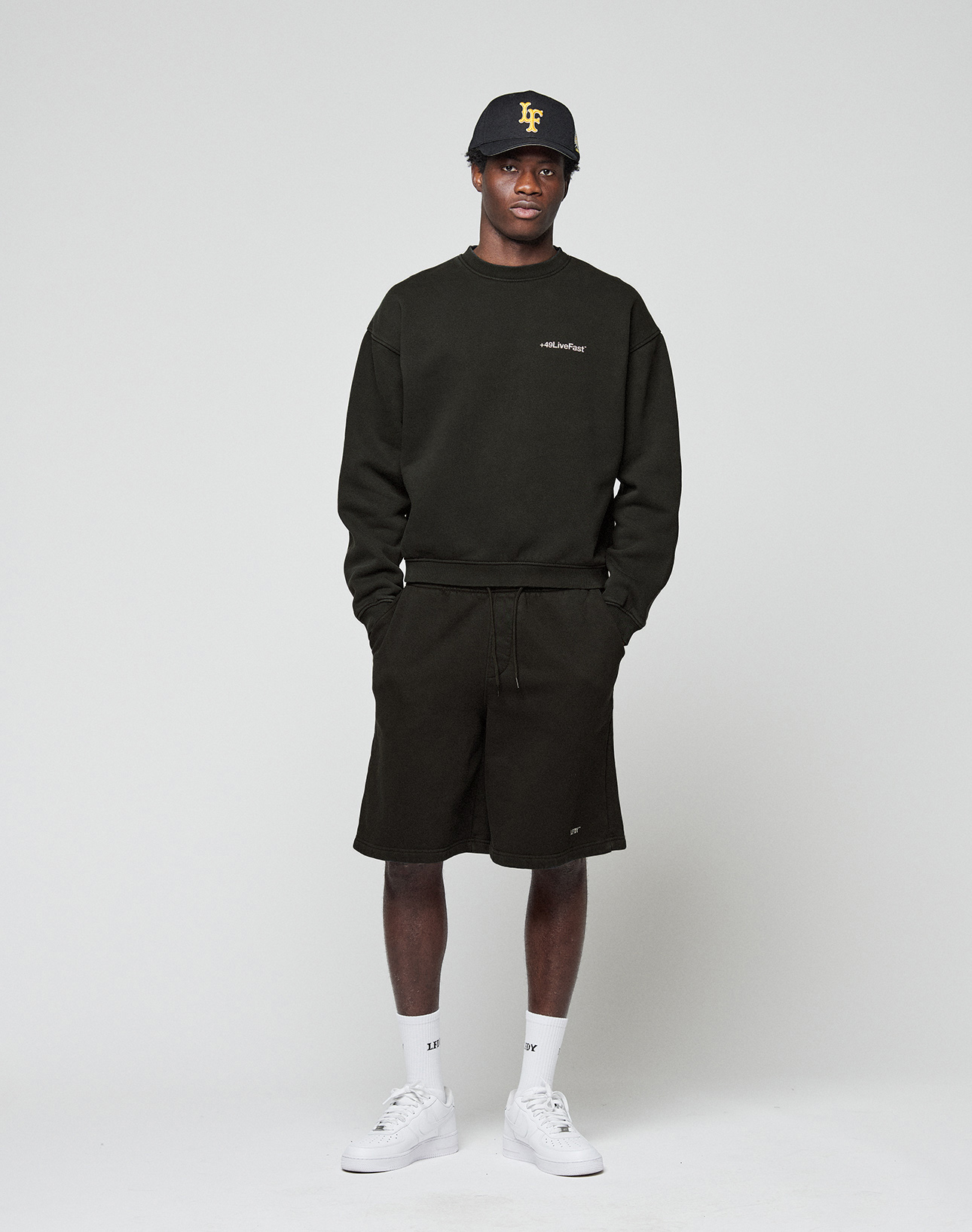 A man faces forward in a LIVE FAST DIE YOUNG Basic +49 Chest Sweater, black shorts, a black cap with yellow lettering, white socks, and white sneakers, set against a plain light background.