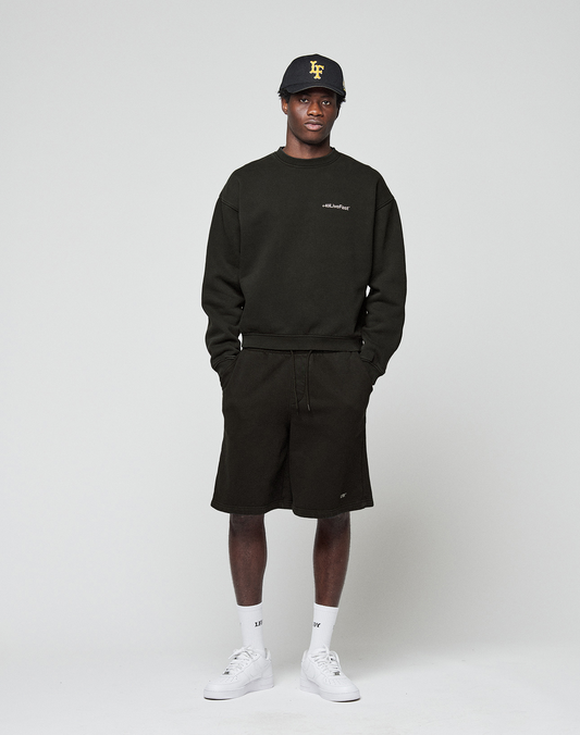 A man faces forward in a LIVE FAST DIE YOUNG Basic +49 Chest Sweater, black shorts, a black cap with yellow lettering, white socks, and white sneakers, set against a plain light background.