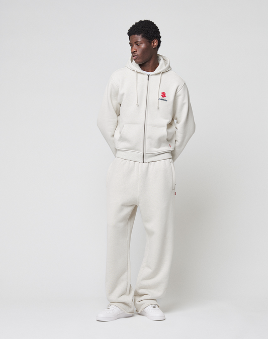 A man stands against a white background wearing LIVE FAST DIE YOUNG's off-white Heavyweight Loungepants and matching 650gsm hoodie with a red chest graphic, paired with white sneakers. His hands are in his pockets as he looks to the side.