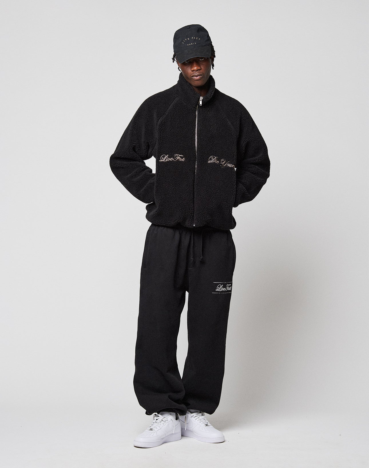 A person stands against a plain background wearing the black LFDY Teddy Jacket by LIVE FAST DIE YOUNG with Raglan sleeves, matching black sweatpants, a black cap, and white sneakers. The jacket and pants feature subtle text details.