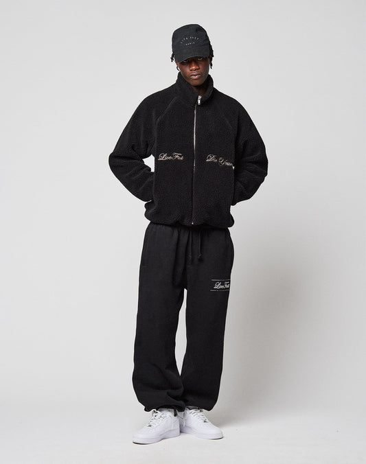 A person stands against a plain background wearing the black LFDY Teddy Jacket by LIVE FAST DIE YOUNG with Raglan sleeves, matching black sweatpants, a black cap, and white sneakers. The jacket and pants feature subtle text details.