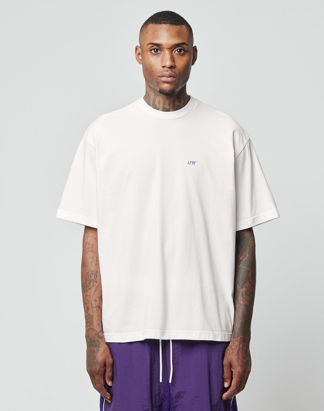 A man with short hair and visible arm tattoos wears the LFDY Originals Tee by LIVE FAST DIE YOUNG in an oversized fit, paired with purple shorts, standing against a plain light gray background and looking directly at the camera.