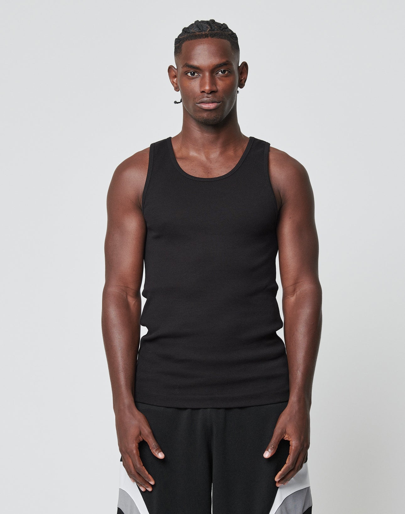 A man stands facing forward against a plain background, wearing the LIVE FAST DIE YOUNG Blank Rib Tank in slim fit black and black-and-white athletic shorts. Both feature Flag Label details and are crafted from Baumwolle. His arms are relaxed at his sides.