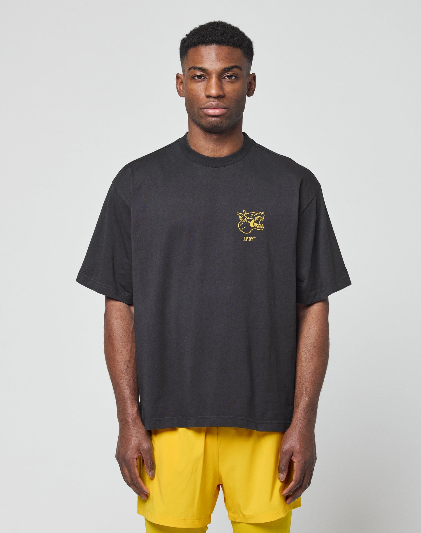 A man faces forward against a plain light gray background, wearing the LIVE FAST DIE YOUNG LFDY Circle Dog Tee in loose oversize fit with a small yellow graphic, paired with yellow shorts.