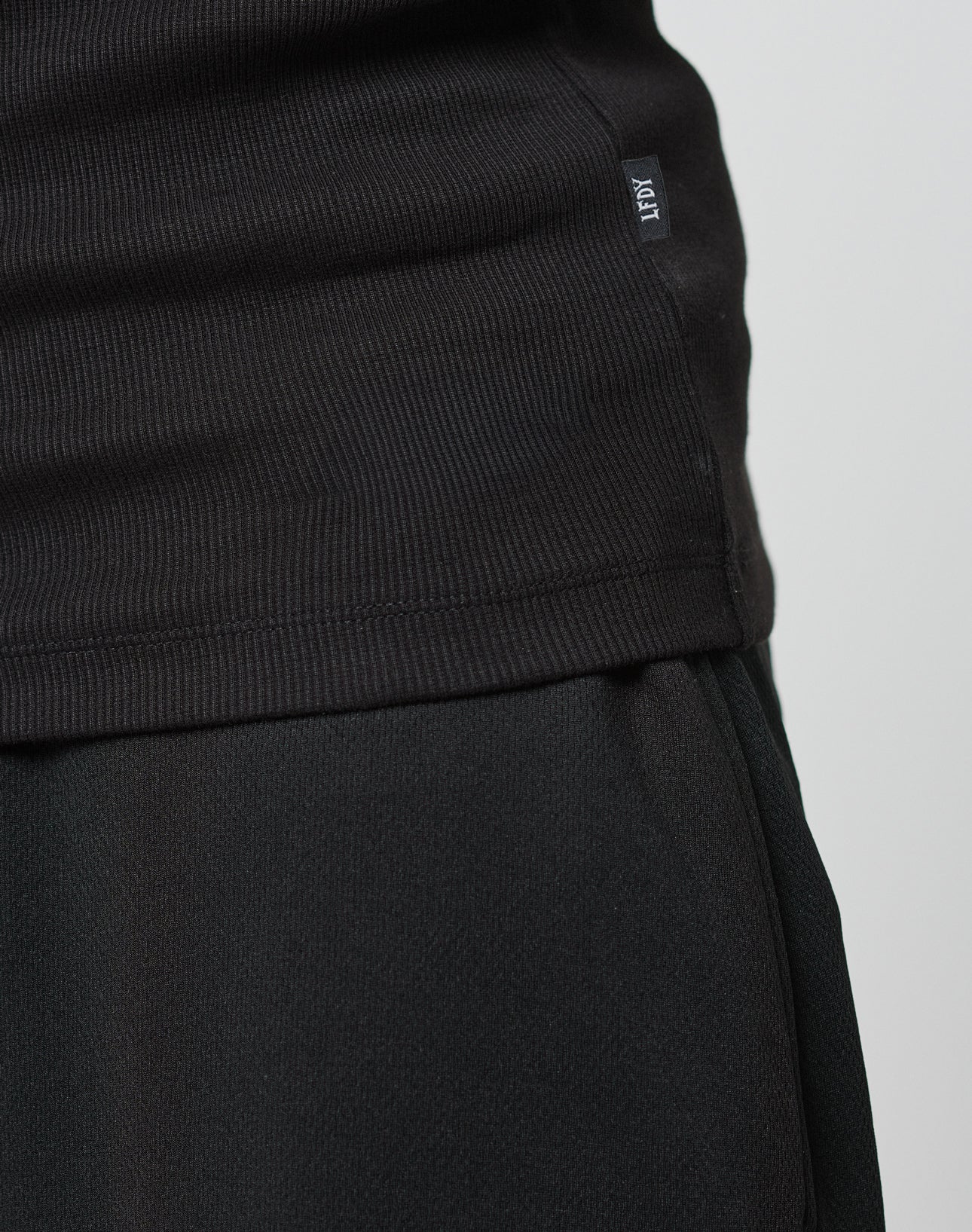 Close-up of a person wearing the LIVE FAST DIE YOUNG Blank Rib Tank, slim fit and ribbed in black, with a Flag Label on the side seam. The neutral background highlights the cotton texture and tank details.