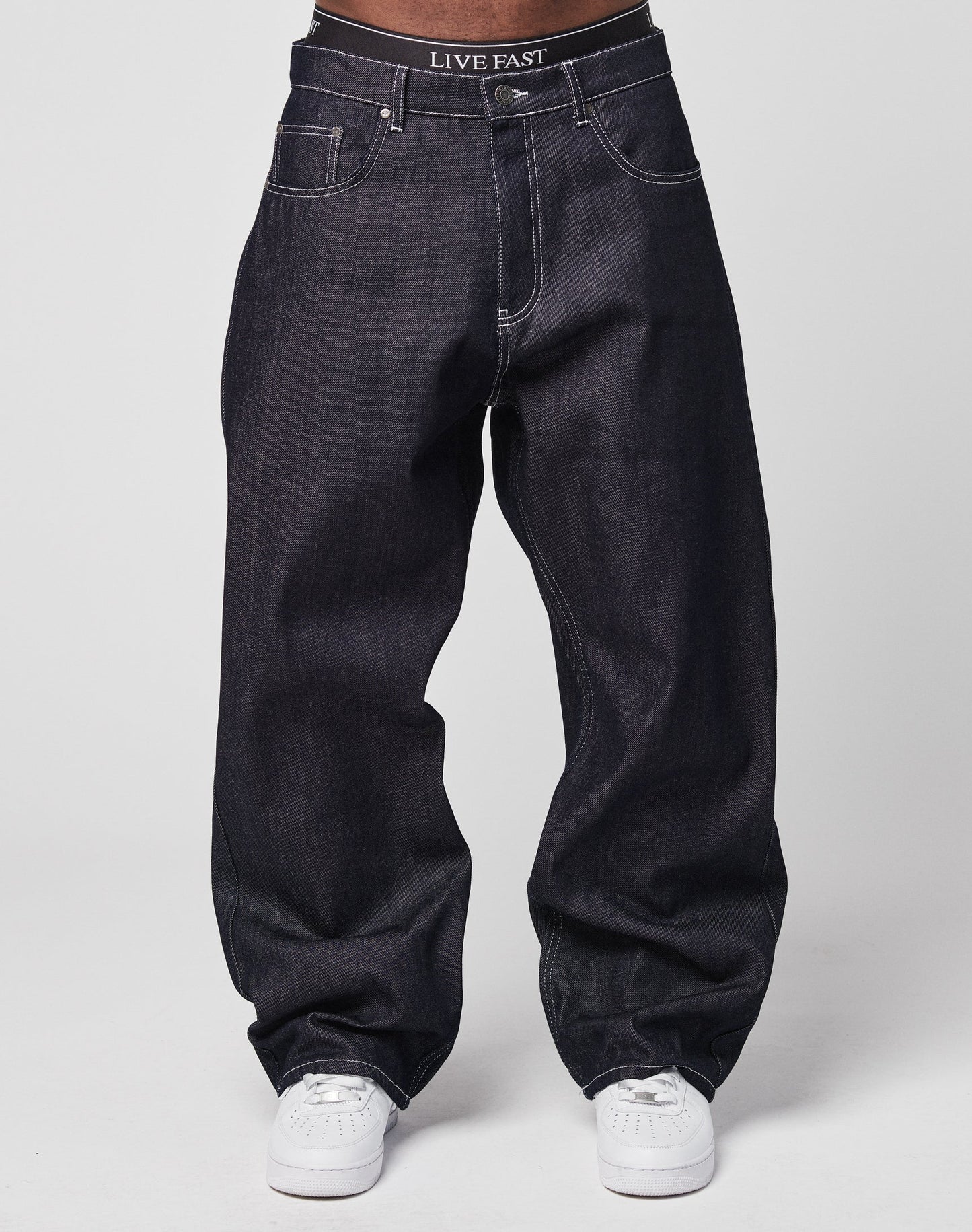 A person in LFDY Baggy Denim with a wide straight leg, 100% cotton, white sneakers, and black LIVE FAST underwear visible above the jeans stands against a plain light background.