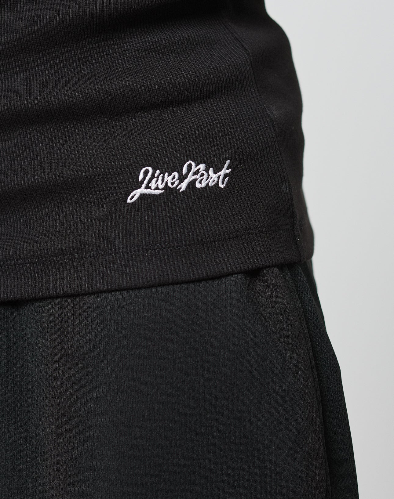 Close-up of a person in slim fit black pants and the LIVE FAST DIE YOUNG Basic Rib Tank, made from cotton and elastane, featuring Live Fast embroidered in white cursive near the hem.