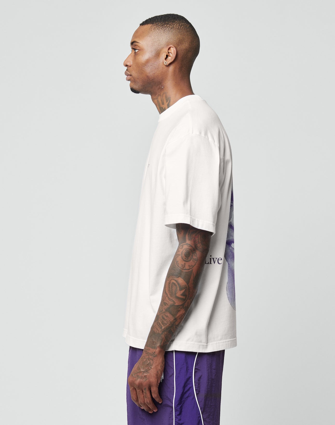 A man with short hair and arm tattoos stands in profile, wearing the oversize fit white LFDY Originals Tee from LIVE FAST DIE YOUNG and purple shorts against a plain light gray background.