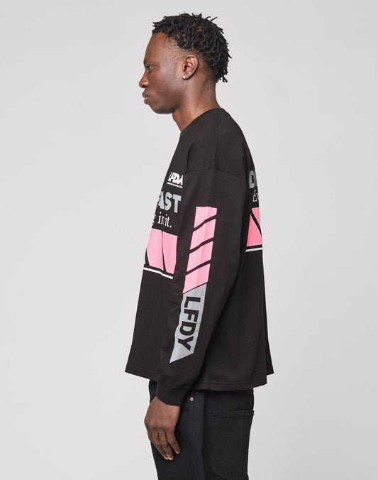 A person with short, twisted hair stands in profile against a plain background, wearing the LFDY Racing Longsleeve by LIVE FAST DIE YOUNG: an oversize black shirt made from 100% Baumwolle with white/pink graphics and reflective prints on sleeves and chest.