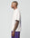 A man with short hair and arm tattoos stands in profile, wearing the oversize fit white LFDY Originals Tee from LIVE FAST DIE YOUNG and purple shorts against a plain light gray background.