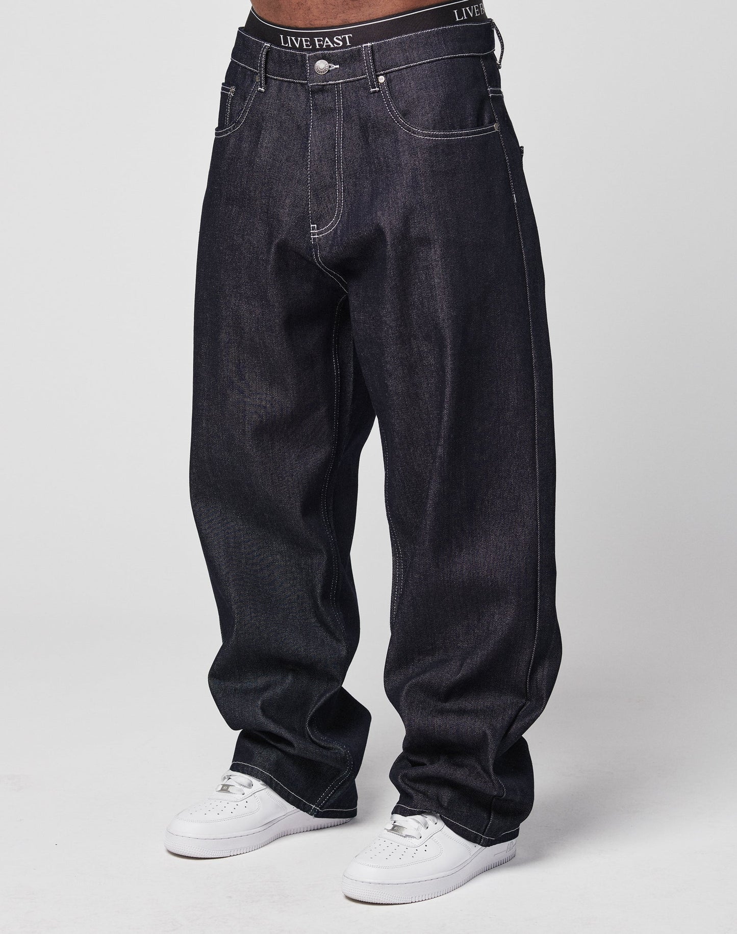 A person wears LFDY Baggy Denim jeans made from 100% cotton with a wide straight leg, white stitching, and a black waistband reading LIVE FAST. Theyre paired with white sneakers against a plain, light background. | model