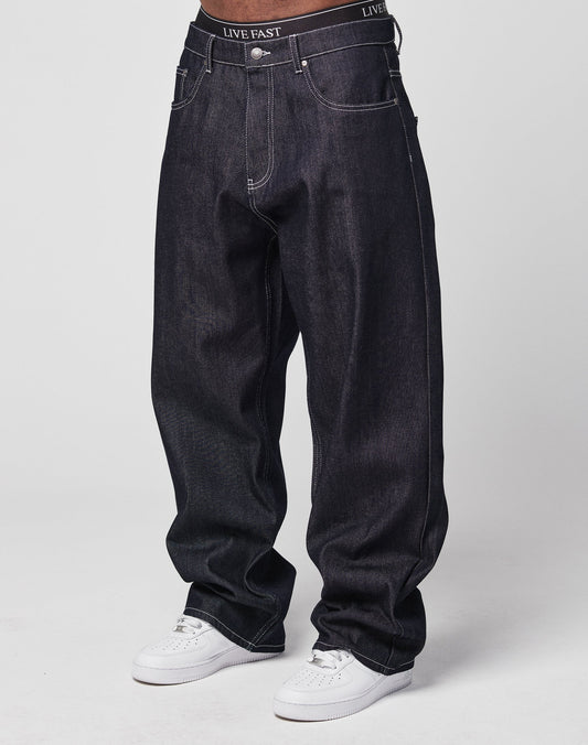 A person wears LFDY Baggy Denim jeans made from 100% cotton with a wide straight leg, white stitching, and a black waistband reading LIVE FAST. Theyre paired with white sneakers against a plain, light background. | model