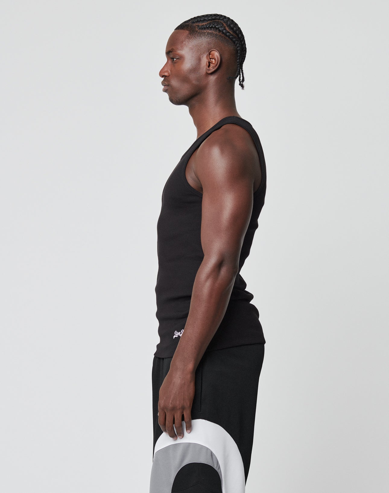 A man with braided hair stands in profile against a plain light background, wearing the LIVE FAST DIE YOUNG Basic Rib Tank and black pants with a white and gray graphic detail on the leg.