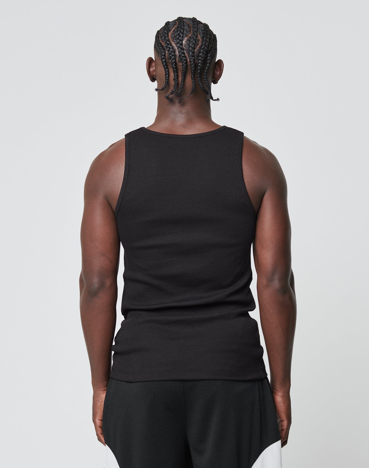 A person with braided hair faces away from the camera, wearing a black LIVE FAST DIE YOUNG Blank Rib Tank made of weicher Baumwolle and black-and-white shorts. A flag label adds a subtle accent against the plain, light background.