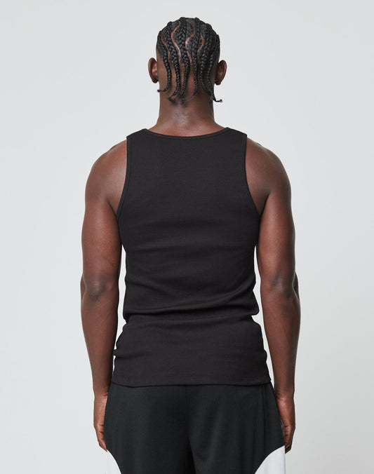 A person with braided hair faces away from the camera, wearing a black LIVE FAST DIE YOUNG Blank Rib Tank made of weicher Baumwolle and black-and-white shorts. A flag label adds a subtle accent against the plain, light background.