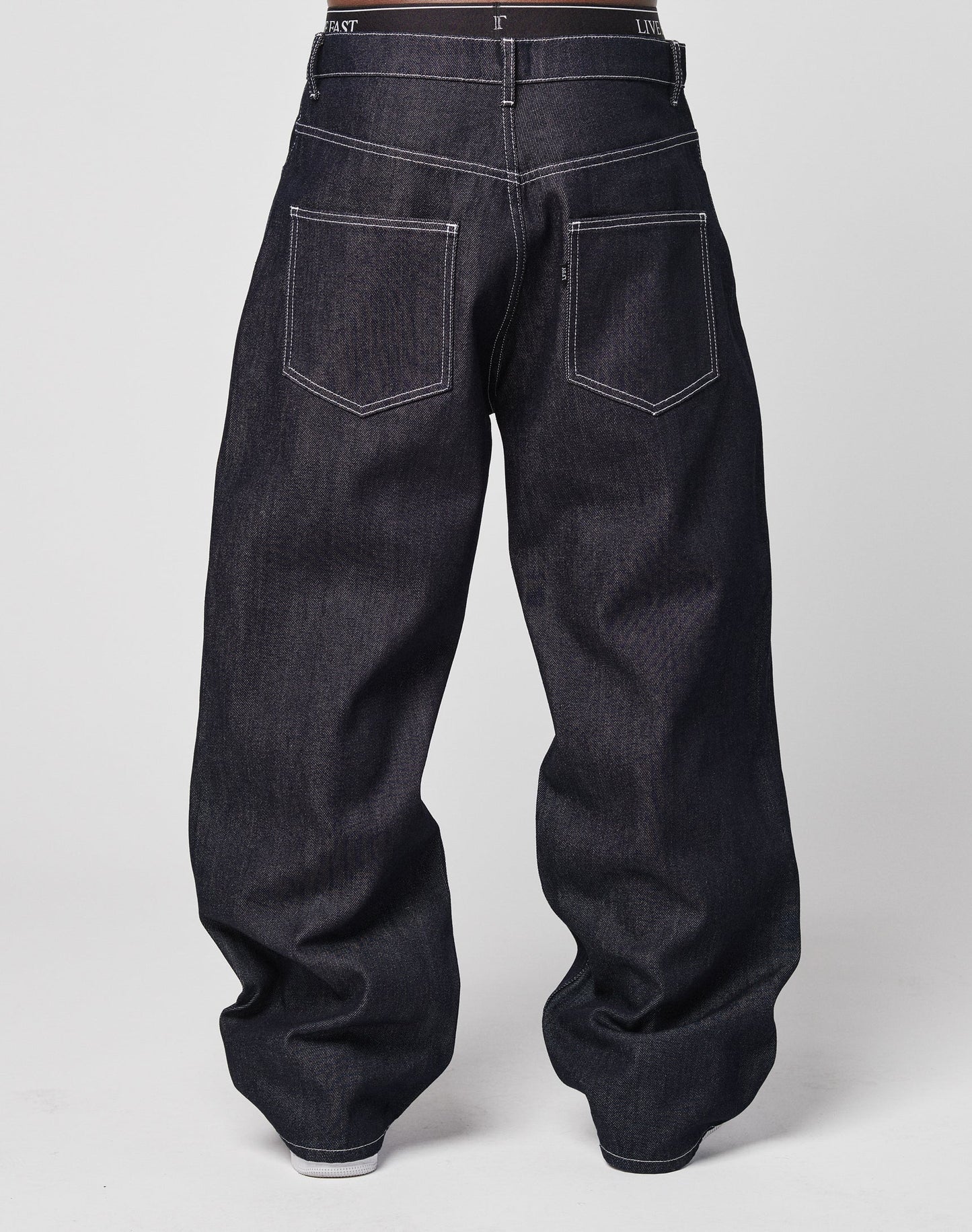A person wearing LFDY Baggy Denim with a wide straight leg and visible white stitching, seen from the back. The loose 100% cotton denim has two large back pockets and covers most of the shoes.