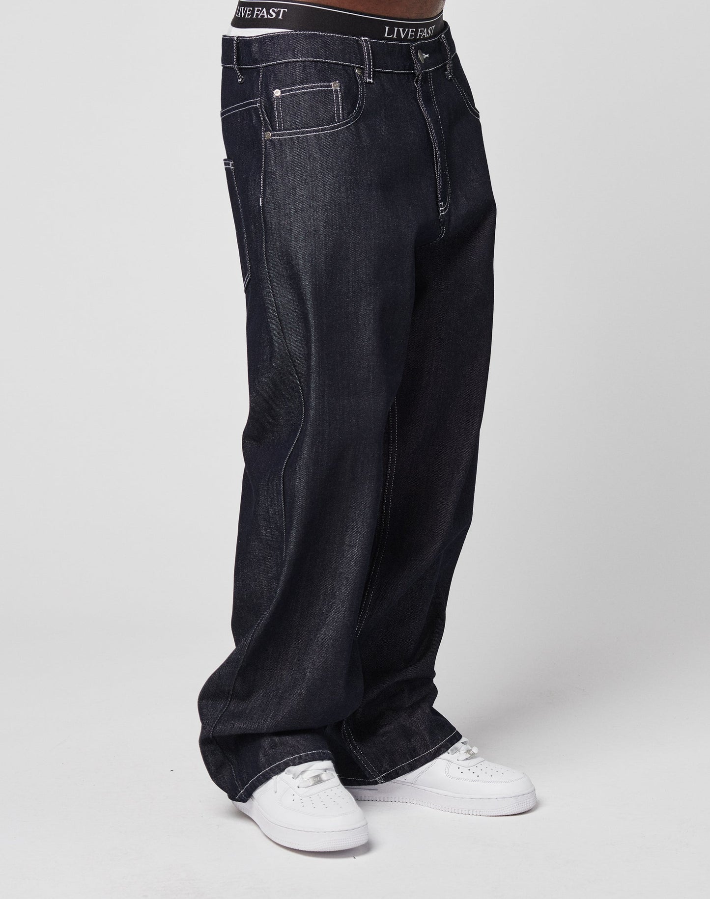 A person wears LIVE FAST DIE YOUNG’s LFDY Baggy Denim with a wide straight leg, white sneakers, and black LIVE FAST underwear showing at the waistband, standing against a plain white background.