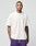 A tattooed man stands facing forward against a light gray background, wearing an oversize plain white LFDY Originals Tee by LIVE FAST DIE YOUNG and purple shorts.
