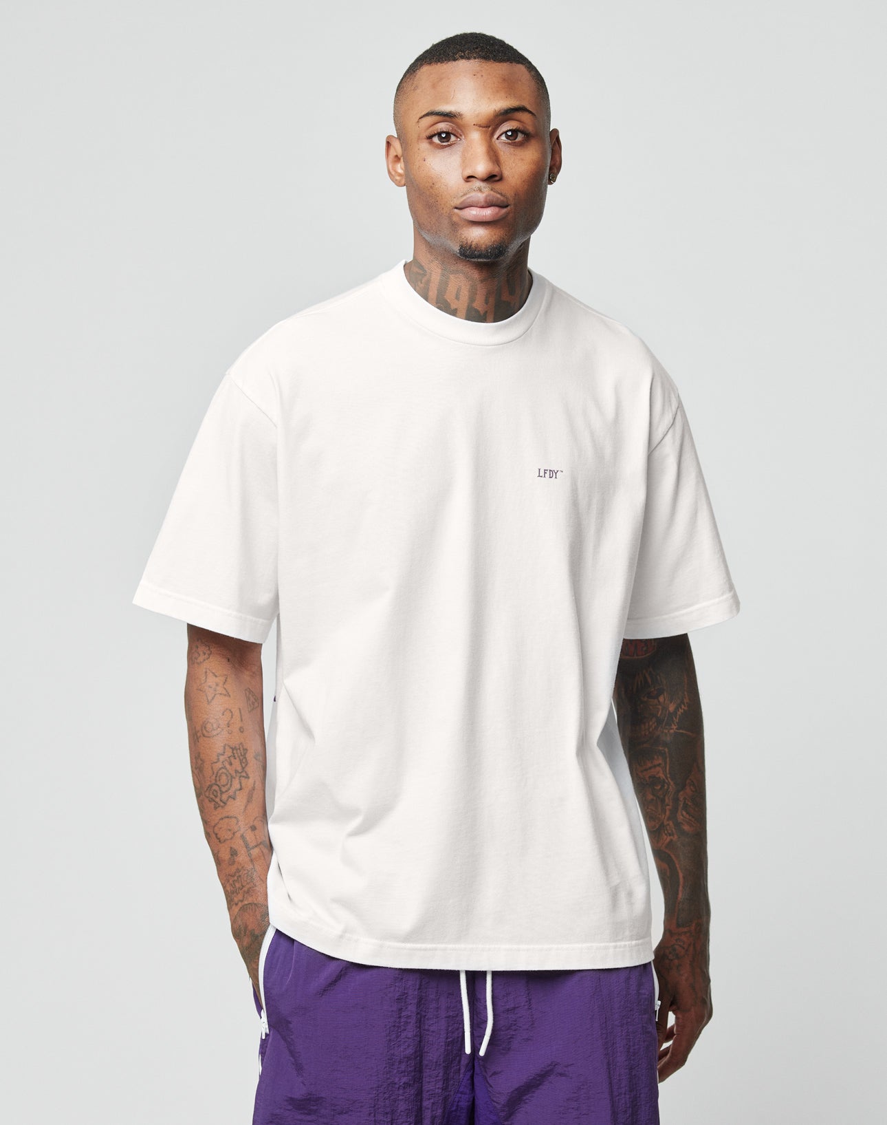 A tattooed man stands facing forward against a light gray background, wearing an oversize plain white LFDY Originals Tee by LIVE FAST DIE YOUNG and purple shorts.