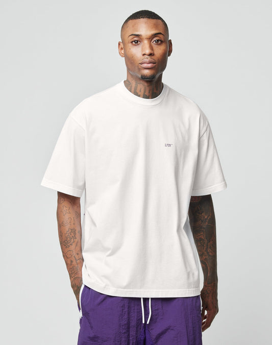 A tattooed man stands facing forward against a light gray background, wearing an oversize plain white LFDY Originals Tee by LIVE FAST DIE YOUNG and purple shorts.