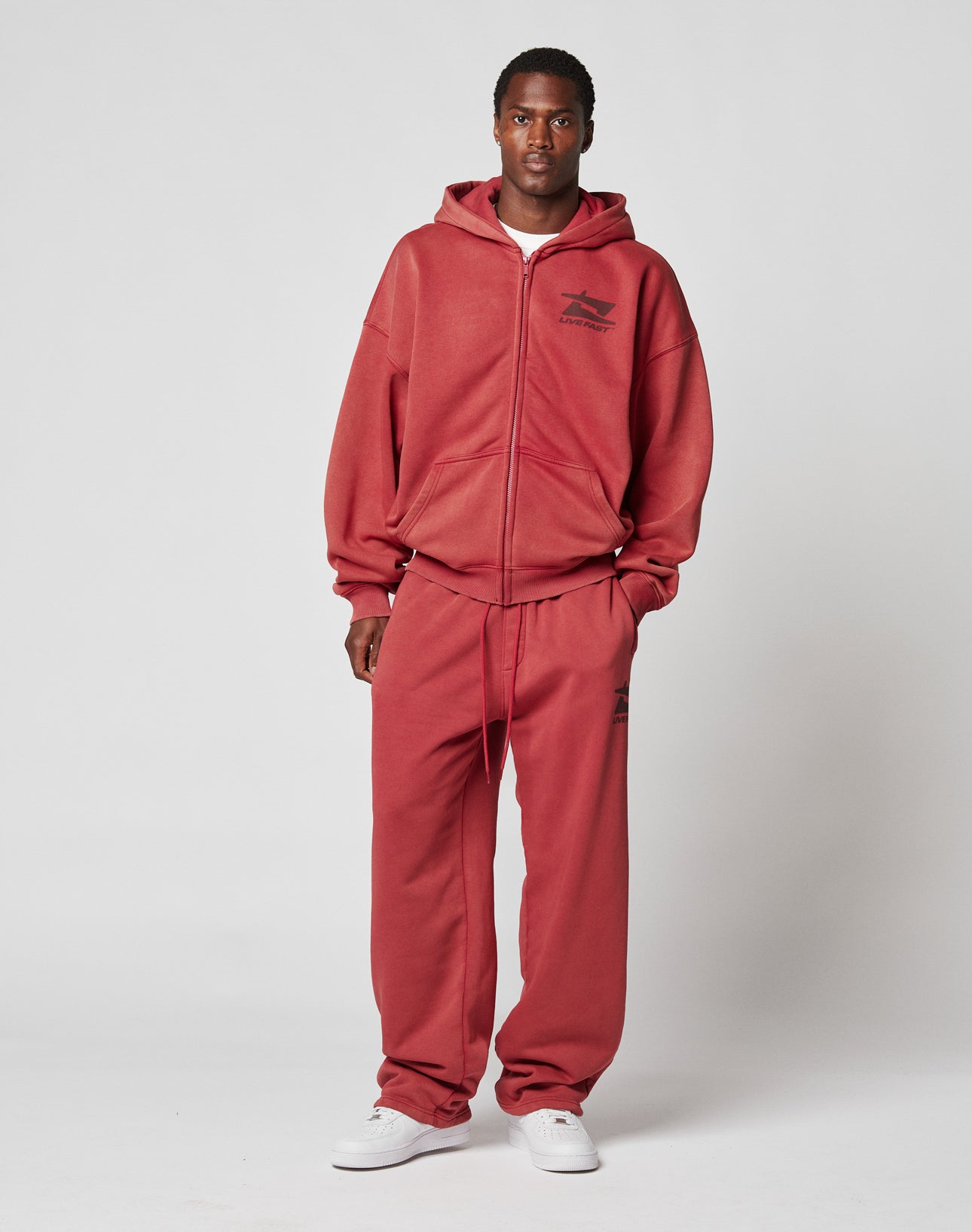 A man faces forward, wearing the LIVE FAST DIE YOUNG Live Fast Sweatsuit Jogger— a loose red zip-up hoodie and matching sweatpants with an elastic waistband and small logo— plus white sneakers, set against a plain light gray background.