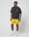 A man wears the LIVE FAST DIE YOUNG LFDY Circle Dog Tee, black and loose with a small yellow logo, paired with yellow shorts, white socks, and white sneakers as he stands against a plain white background looking to his left.