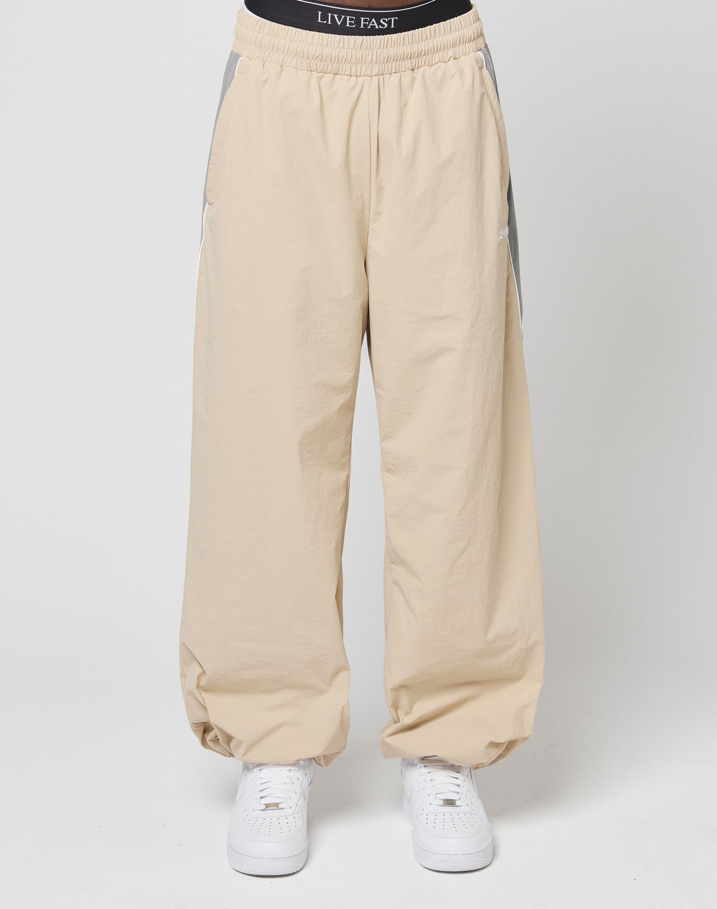 A person models LIVE FAST DIE YOUNGs Basic Nylon Trackpants in wide fit beige with gray side panels, an elastik-kordelzug waist, and a visible black LIVE FAST waistband, paired with white sneakers against a plain white background.