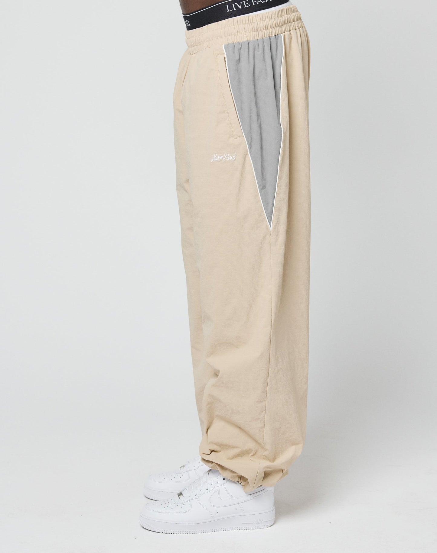 A person is wearing LIVE FAST DIE YOUNG Basic Nylon Trackpants in beige with a light gray side panel, Reißverschluss Seitentaschen, and an Elastik-Kordelzug, paired with white sneakers and standing sideways against a plain white background.