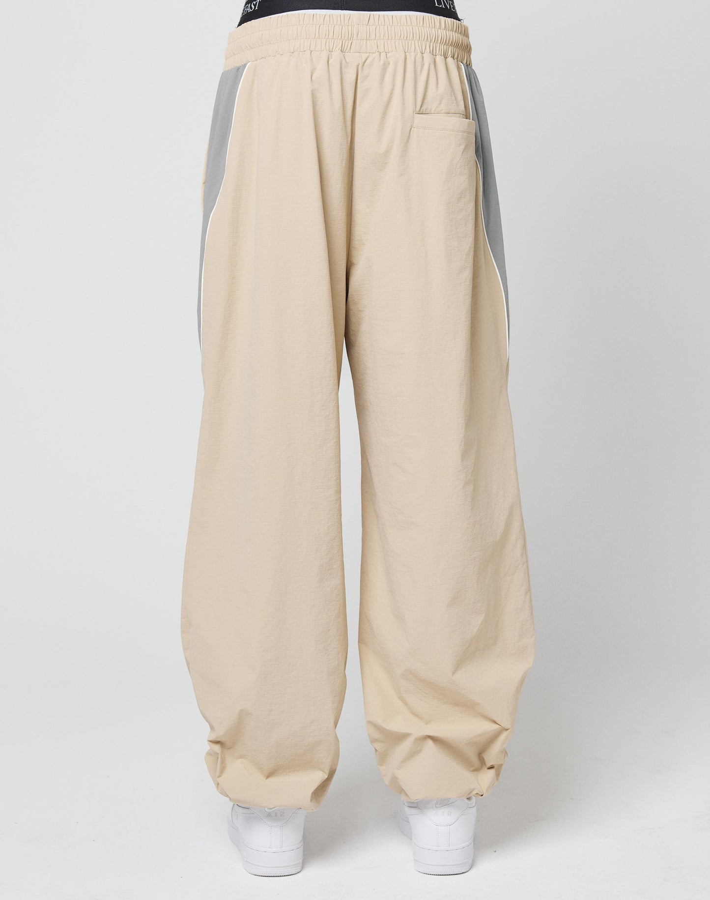 A person wearing LIVE FAST DIE YOUNG Basic Nylon Trackpants in wide fit, beige with gray side panels, elastic drawstring waist, zip side pockets, a back pocket, and white sneakers stands against a plain white background.
