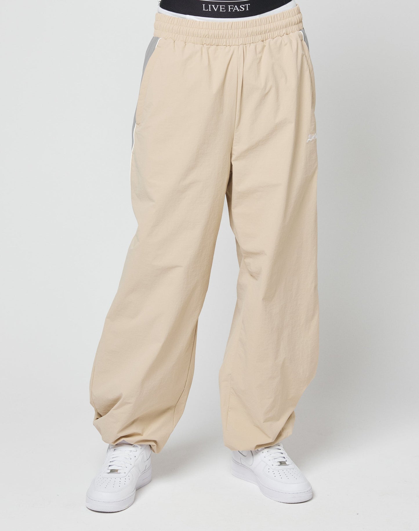 A person wears beige Basic Nylon Trackpants from LIVE FAST DIE YOUNG, featuring an elastic waistband and white sneakers. The black LIVE FAST DIE YOUNG underwear’s white logo is visible above the pants. Background is plain and light-colored.