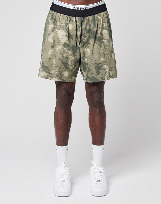 Someone wears LFDY Performance Running Shorts by LIVE FAST DIE YOUNG—green camouflage with a black waistband reading LIVE FAST and a reflective logo print—paired with white socks and sneakers, standing against a plain light background.
