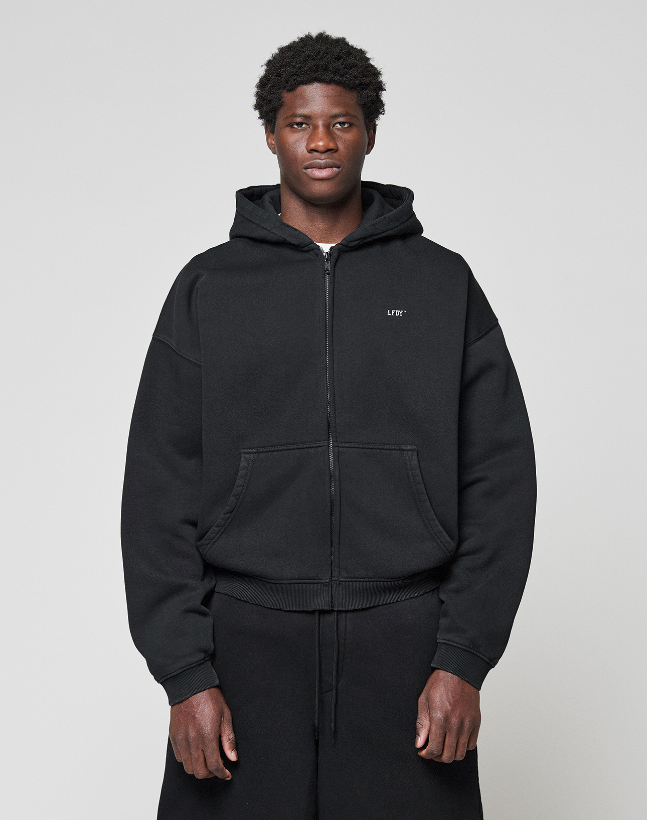 A young man stands against a plain background, wearing the LIVE FAST DIE YOUNG Basic Evidence Hooded Zip in black with subtle branding on the left chest and matching black pants. His hands are by his sides as he looks straight at the camera.