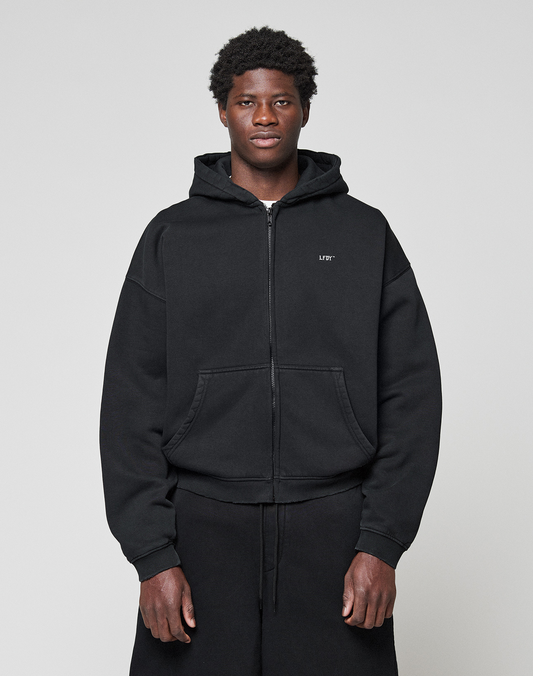 A young man stands against a plain background, wearing the LIVE FAST DIE YOUNG Basic Evidence Hooded Zip in black with subtle branding on the left chest and matching black pants. His hands are by his sides as he looks straight at the camera.