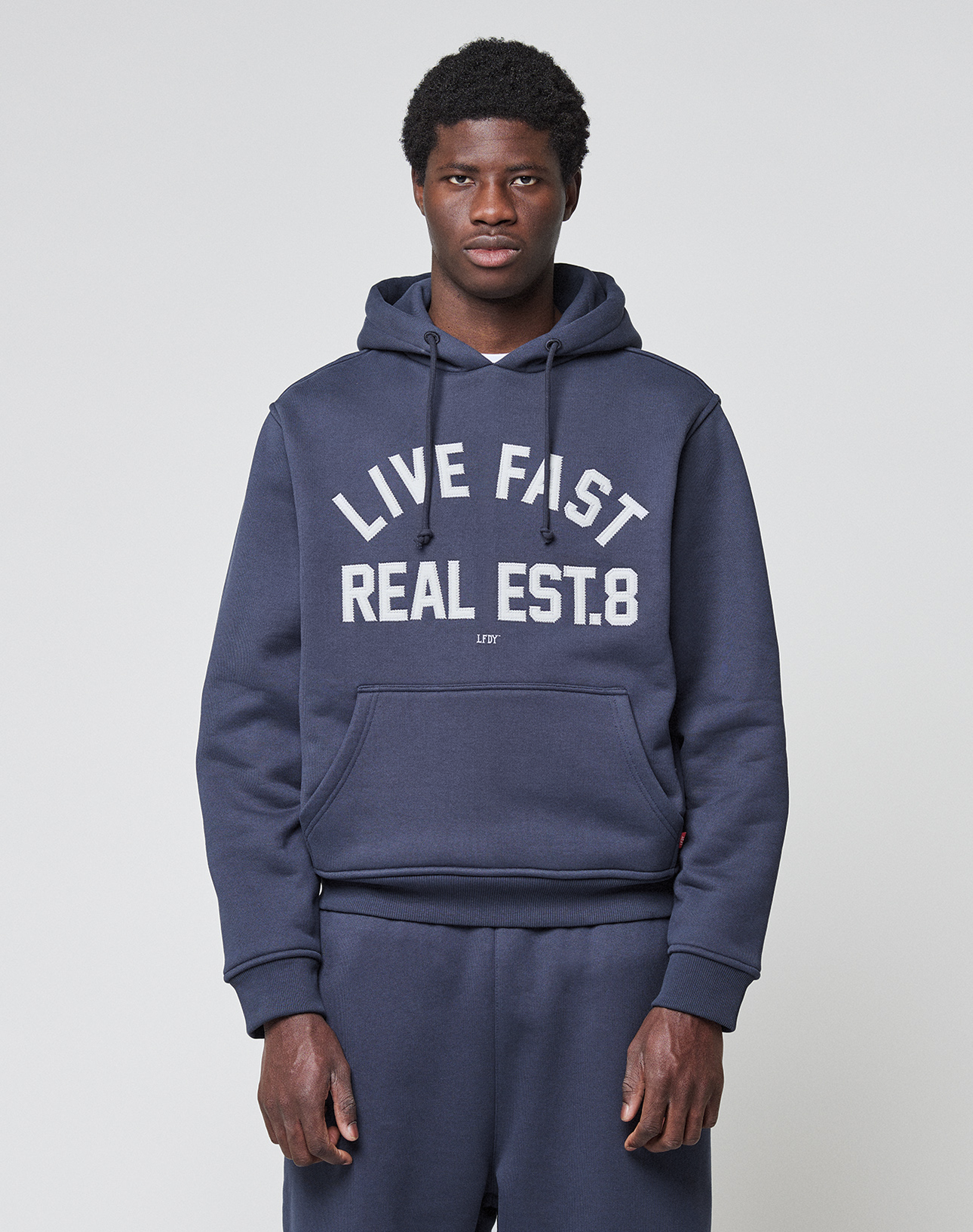 A man faces forward in a navy LFDY Heavyweight Lounge Hooded sweatshirt and matching sweatpants by LIVE FAST DIE YOUNG. The Regular Fit 100% Baumwolle hoodie displays "LIVE FAST REAL EST.8" in large white letters on a plain light background.