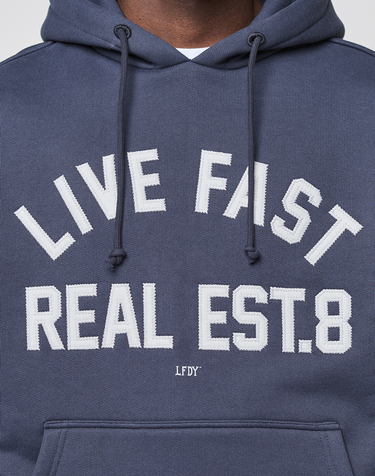 A person wears the LIVE FAST DIE YOUNG LFDY Heavyweight Lounge Hooded in dark blue with white "LIVE FAST REAL EST. 8" text, small LFDY lettering below, made from 100% Baumwolle, regular fit; only the upper chest and part of the face are visible.