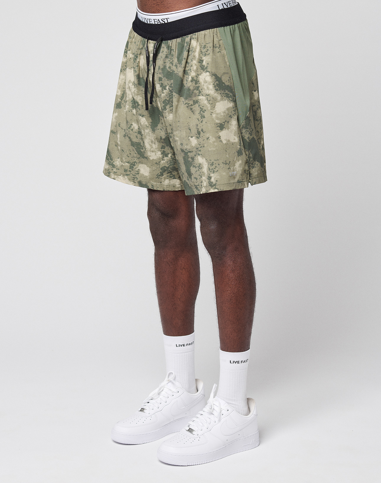 A person wearing green camo LFDY Performance Running Shorts by LIVE FAST DIE YOUNG, featuring a white LIVE FAST waistband, white socks, and white Nike sneakers stands against a plain white background. Only the lower body is visible. | model
