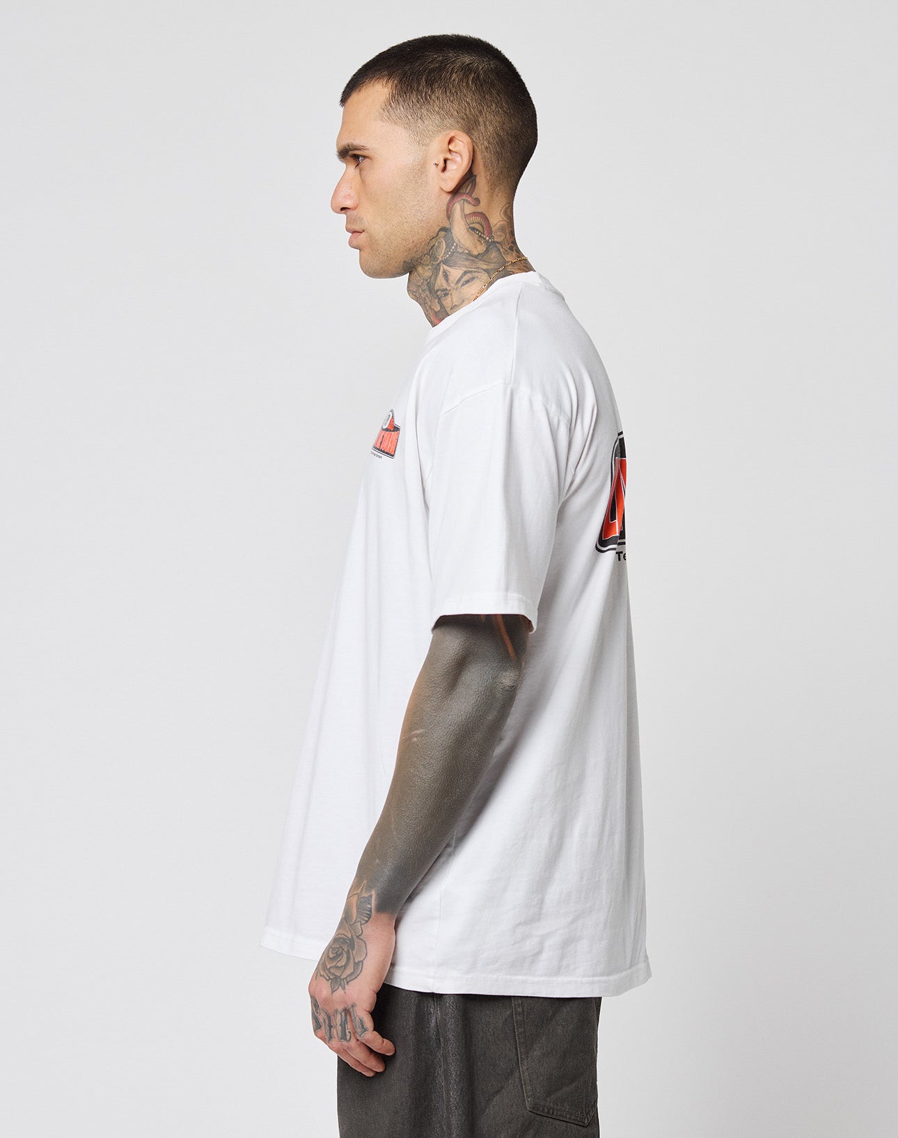 A tattooed individual stands in profile, wearing the LIVE FAST DIE YOUNG Live Fast 2013 Tee—crafted from 100% Baumwolle—and dark pants with subtle topstitching, set against a plain light background.
