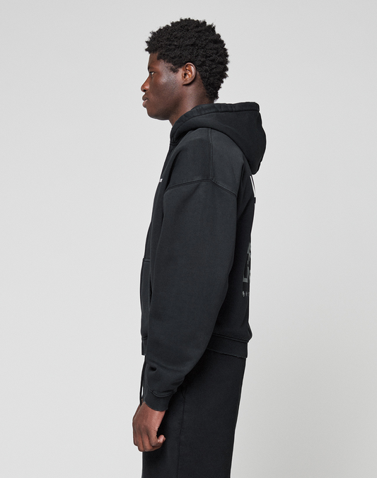 A person with short curly hair stands in profile facing left, wearing a black LIVE FAST DIE YOUNG Basic Evidence Hooded Zip and black pants with subtle branding, against a plain light gray background.